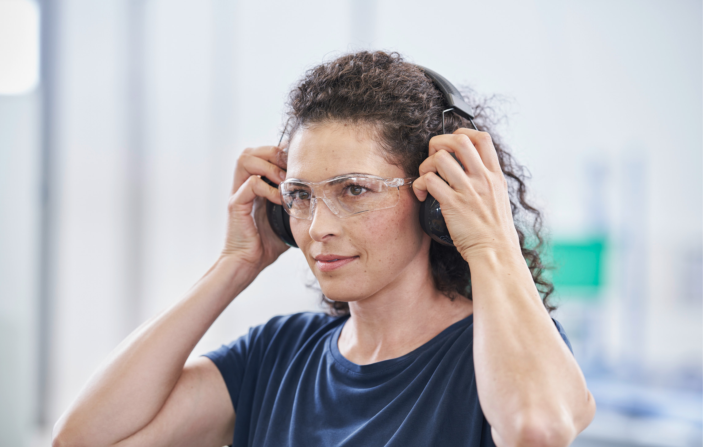 Eine Frau mit gelocktem, braunem Haar und durchsichtiger Schutzbrille zieht sich einen Gehörschutz über den Kopf