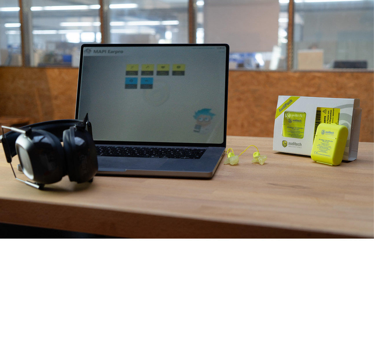 Abbildung eines Laptops auf einer Arbeitsplatte. Links ein Kapselgehörschutz, rechts ein angepasster Gehörschutz mit Verpackung