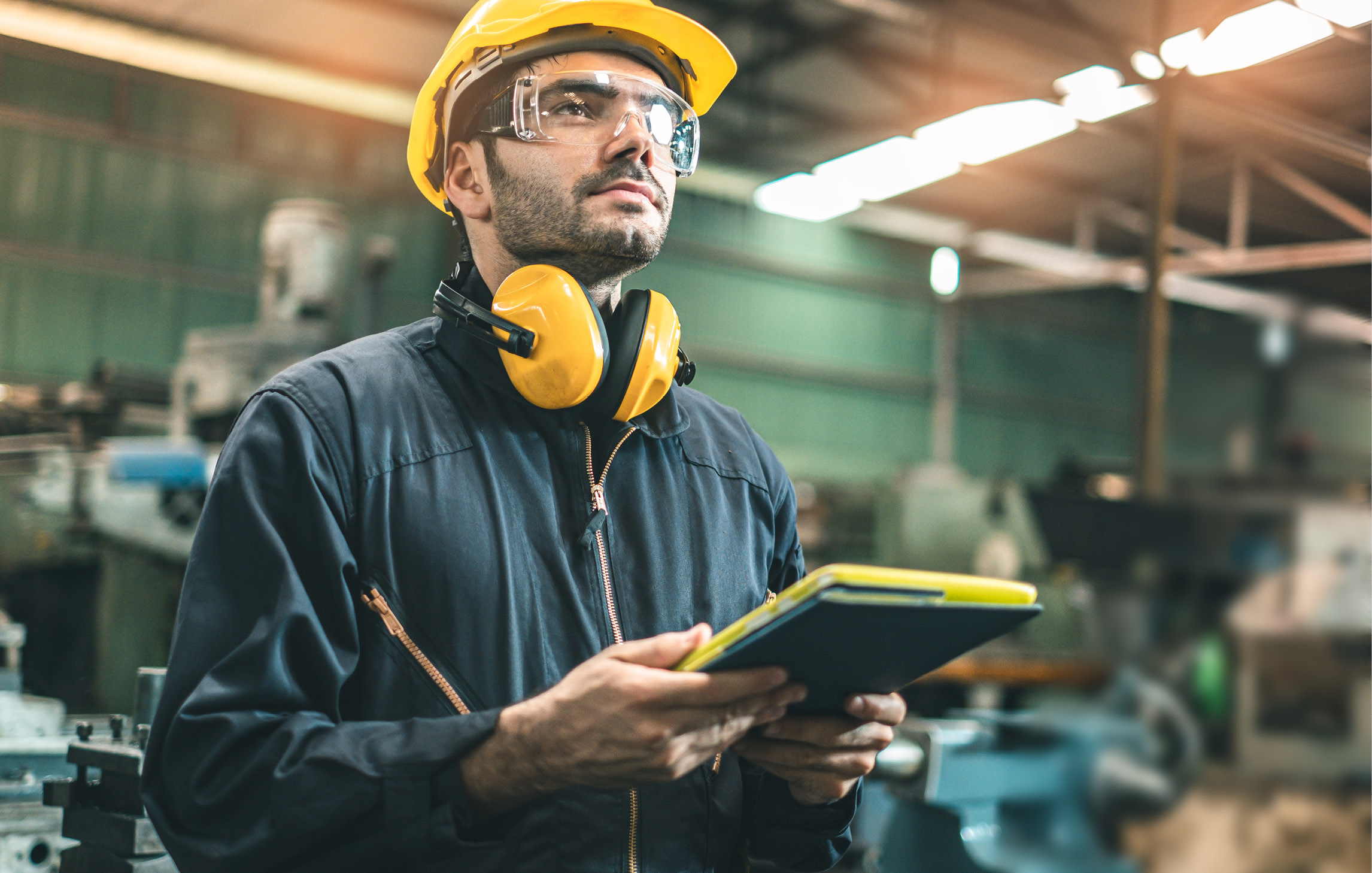Ein Bauarbeiter steht mit einem Klemmbrett in einer Werkstatt. Er trägt einen Overall, Schutzbrille, Helm und Kapselgehörschutz um den Hals