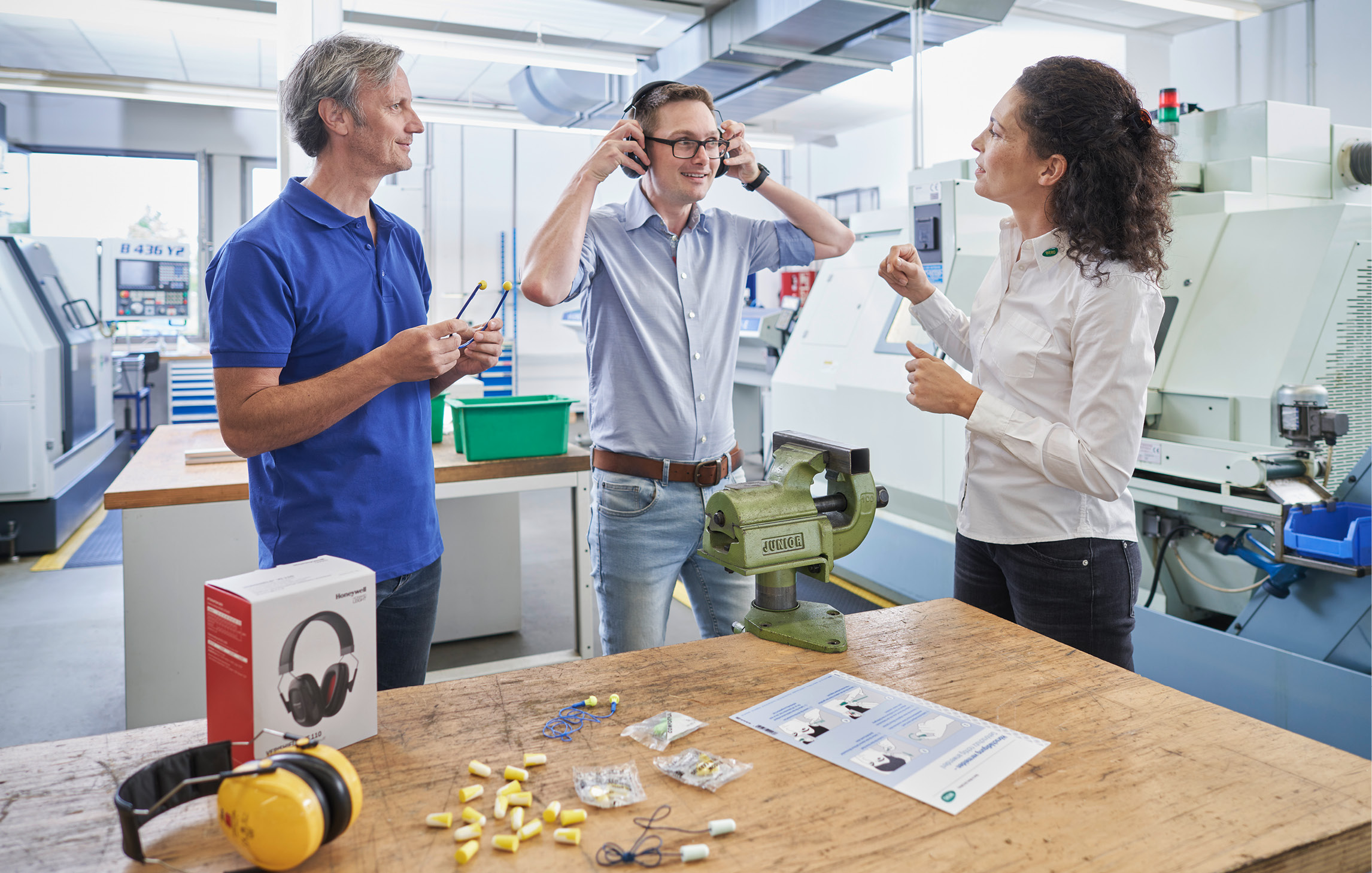 Zwei Personen stehen in einer Werkstatt um eine Arbeitsplatte und testen mit einer WERO Mitarbeitenden verschieden Sorten Gehörschutz