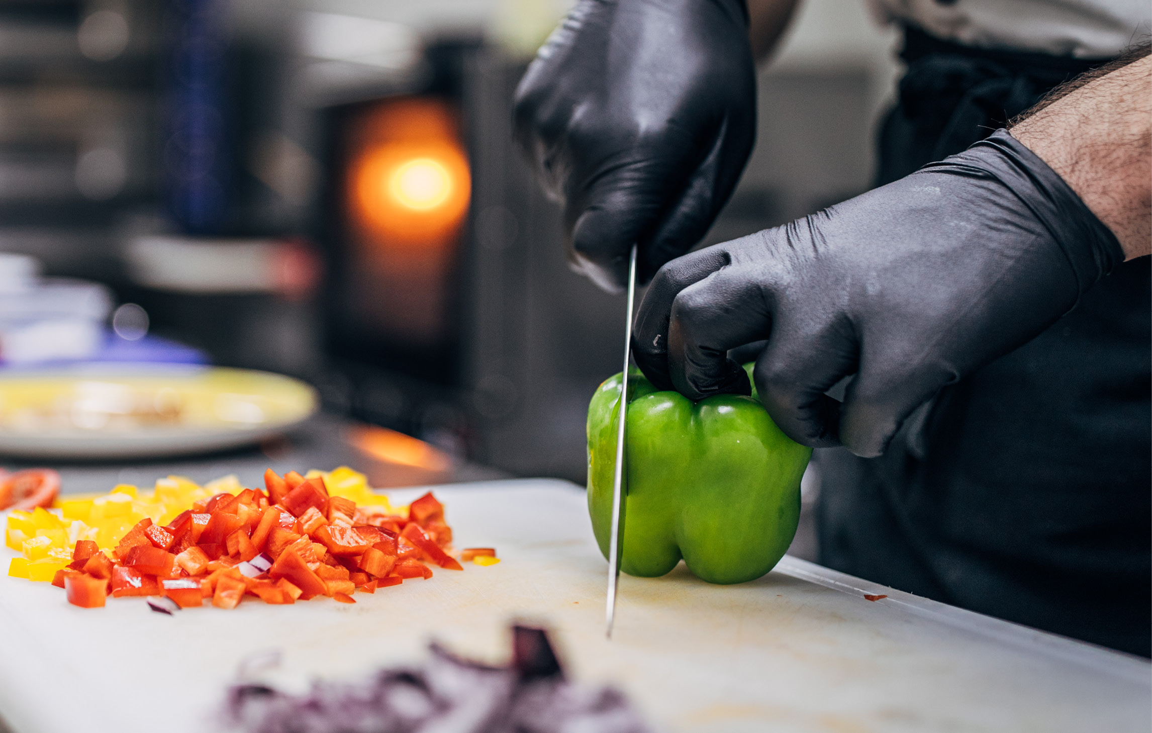 Situation in einer Großküche. Ein Mann mit schwarzen Einmalhandschuhen schneidet eine grüne Paprika. Auf dem Schneidebrett liegt weiteres, geschnittenes Gemüse