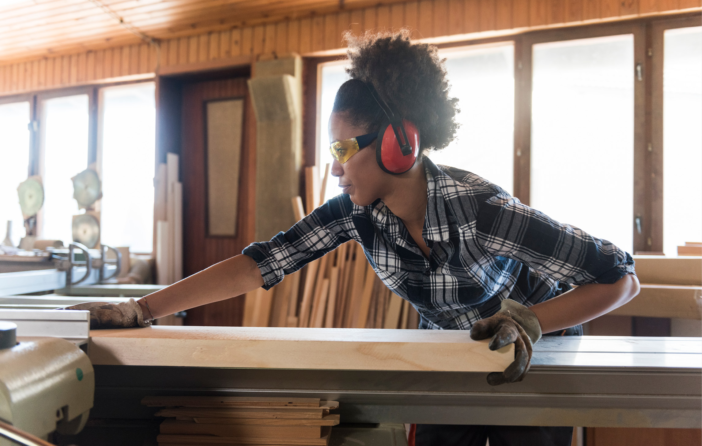 Eine Frau schneidet in einer Werkstatt ein großes Stück Holz zurecht. Sie trägt einen Kapselgehörschutz und Arbeitshandschuhe