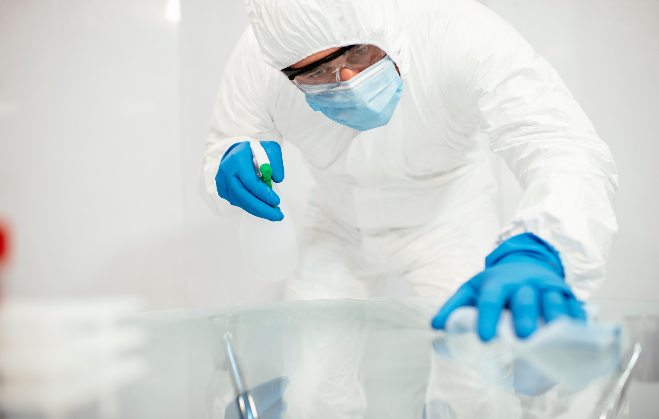 Ein Mann in einem weißen Ganzkörper-Anzug, mit Maske, Schutzbrille und blauen Einmalhandschuhen desinfiziert eine Oberfläche